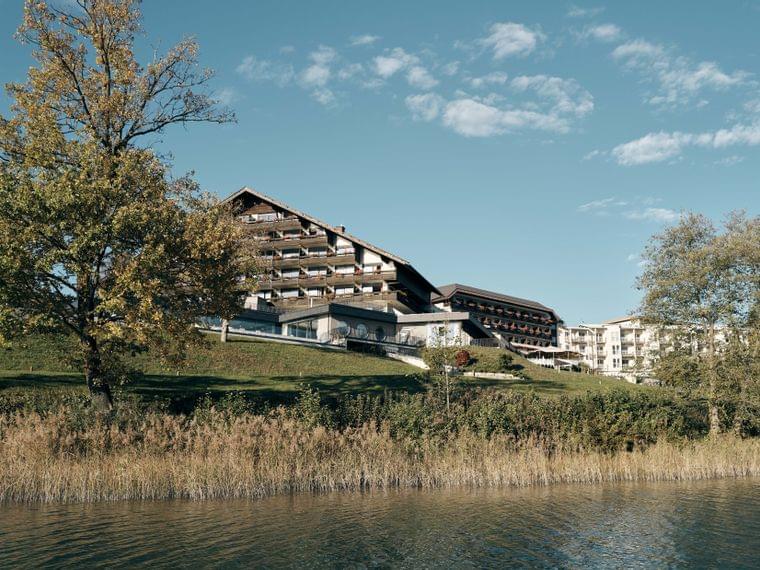 Hotel am See mit Holzbalkonen, umgeben von Bäumen unter klarem blauem Himmel