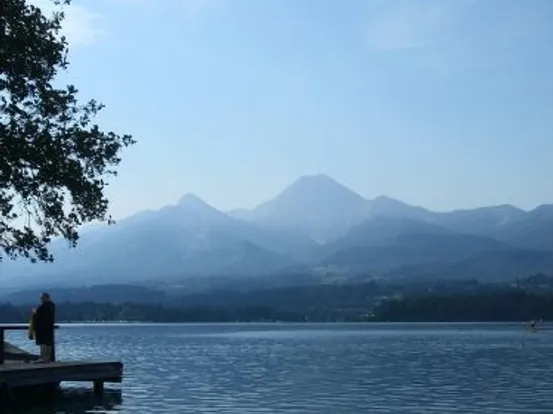 Person steht auf Holzsteg an ruhigem See mit Bergkette im Hintergrund