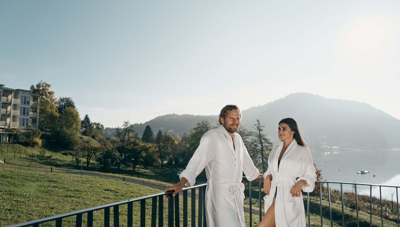 Couple in white bathrobes relaxing on spa hotel balcony with lake and mountain view