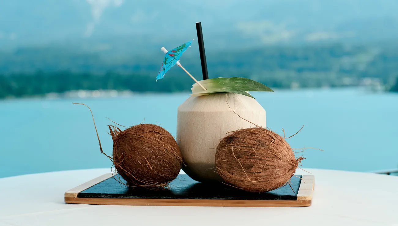 Frisches Kokosgetränk mit Schirmchen und zwei Kokosnüssen vor See- und Bergkulisse