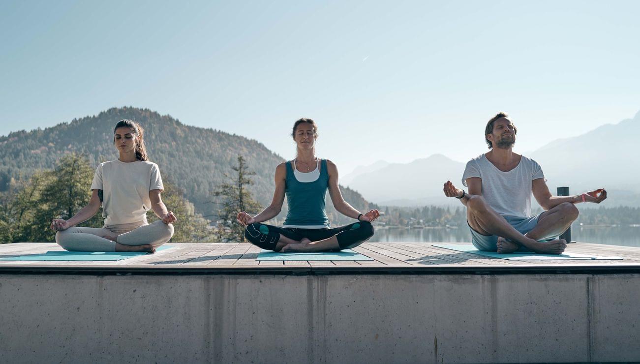 Three people meditating on yoga mats outdoors with mountain lake view