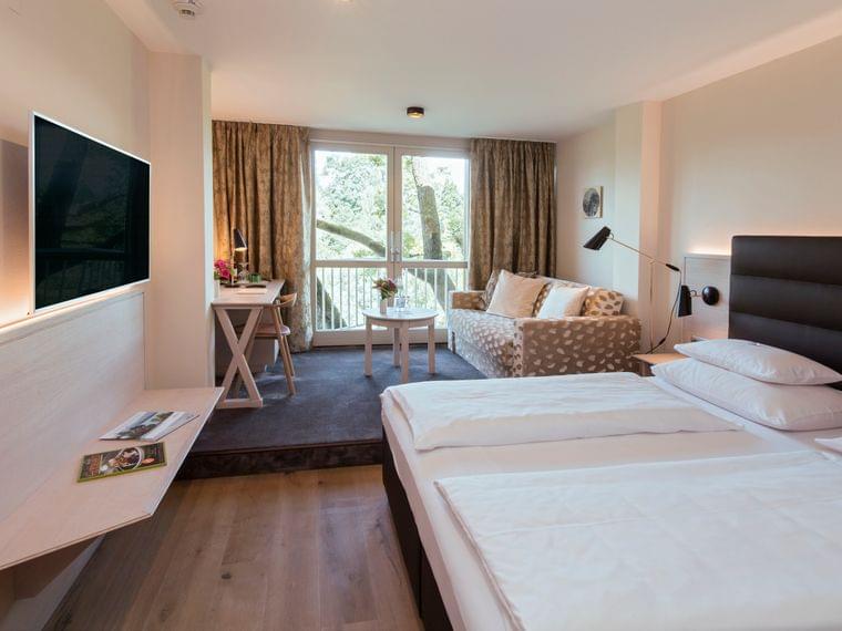 Modernes Hotelzimmer mit Doppelbett, Sofa, Schreibtisch und Balkon mit Waldblick