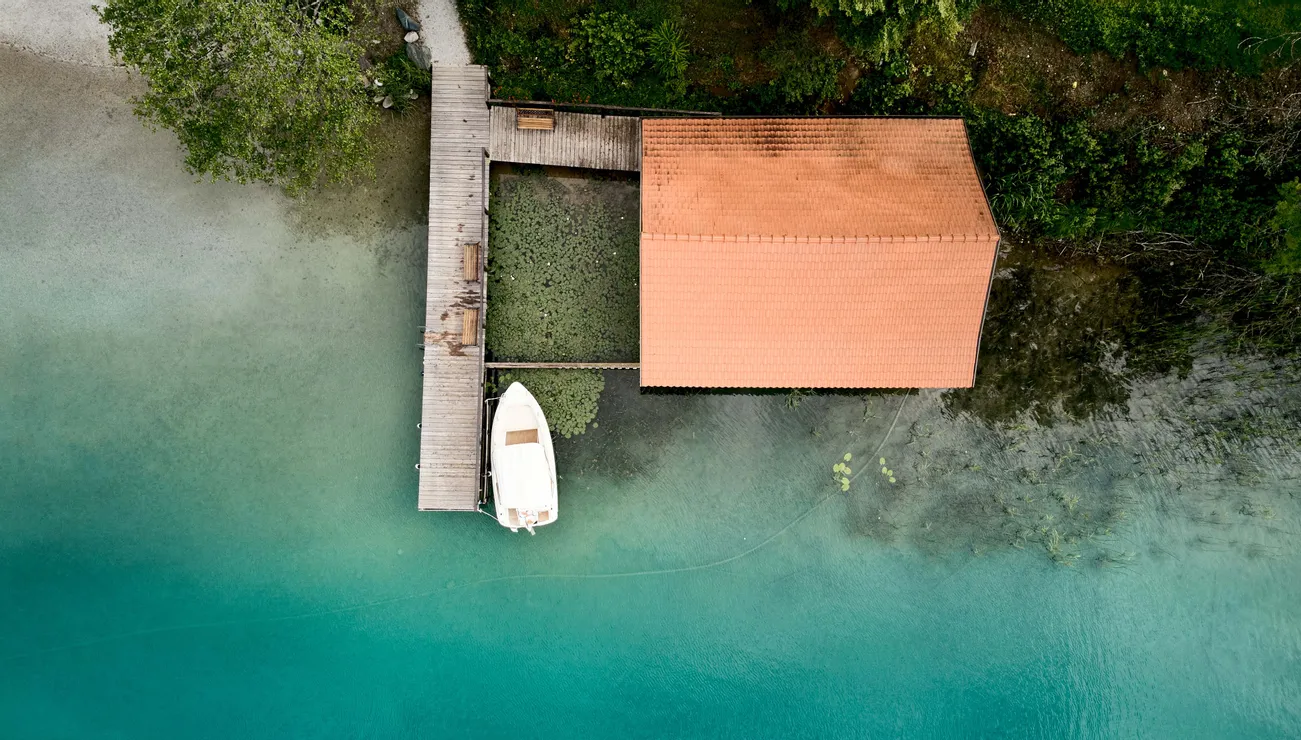Aerial view of a boat docked at a wooden pier next to a boathouse on turquoise lake
