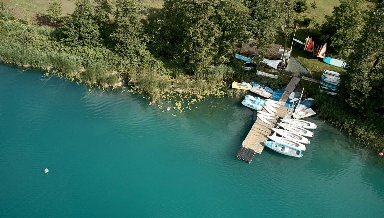 Piccolo molo con pedalò e barche a vela ormeggiati su lago turchese vicino alla riva alberata