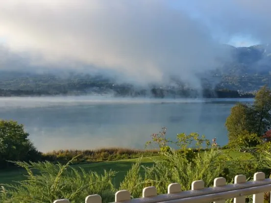 Nebliger Bergsee-Ausblick vom Balkon mit üppigem Grün und Dunst über dem Wasser