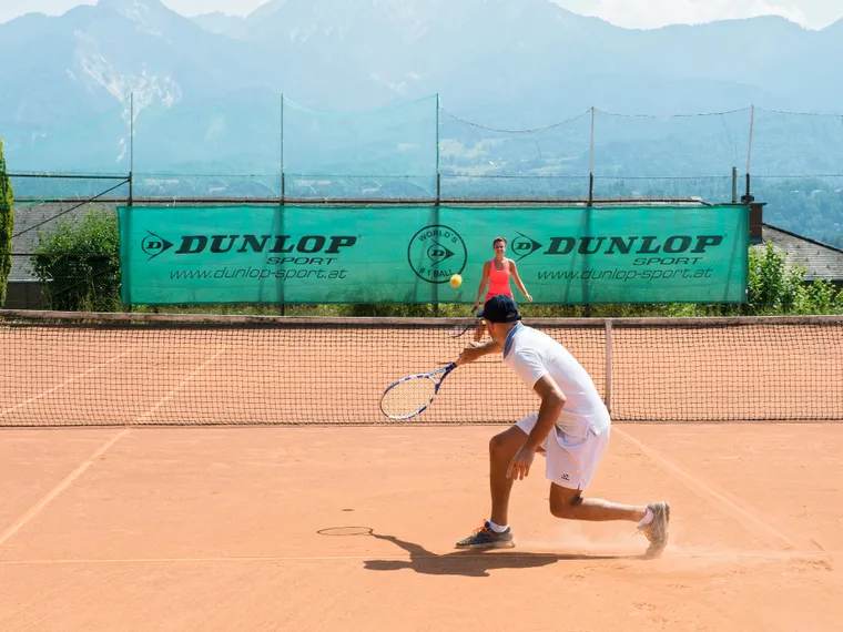 Mann schlägt Tennisball auf Sandplatz mit Bergen im Hintergrund
