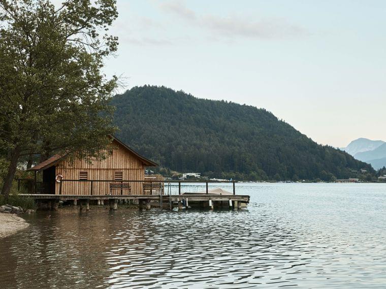 Holzbootshaus am ruhigen Seeufer mit bewaldetem Hügel und Bergen im Hintergrund