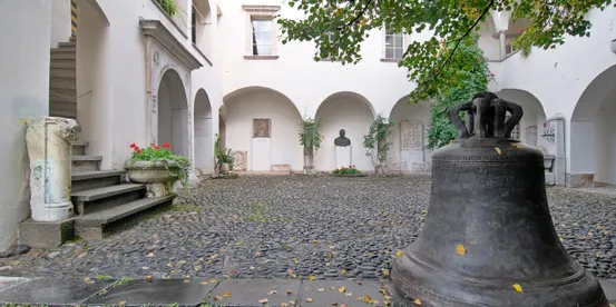 Historischer Innenhof mit Kopfsteinpflaster, großer Bronzeglocke, Arkaden und klassischen Skulpturen