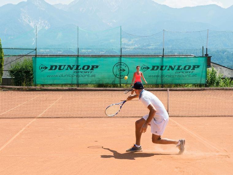 Mann schlägt Tennisball auf Sandplatz mit Bergen im Hintergrund