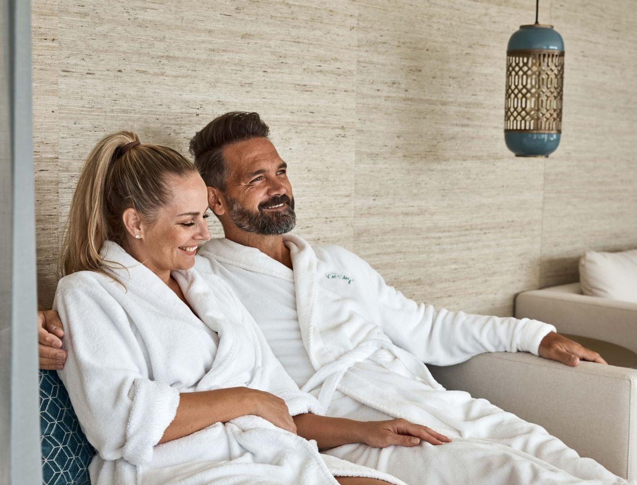Smiling couple in white bathrobes relaxing on a spa lounge sofa