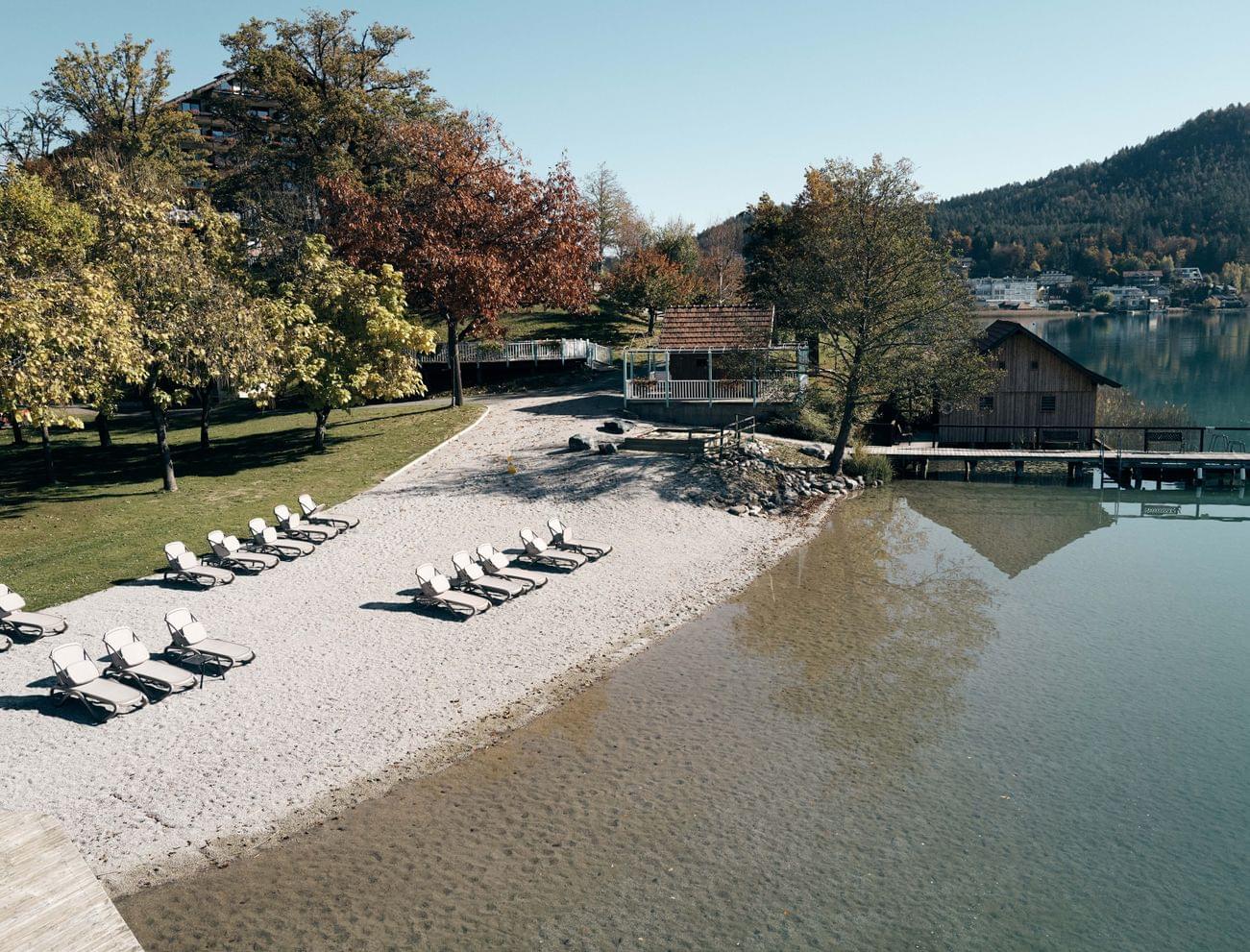 Leere Sonnenliegen an einem ruhigen Seeufer mit klarem Wasser und Herbstbäumen