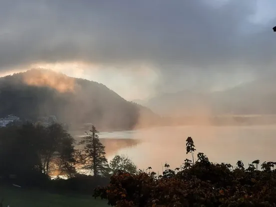 Nebliger See im Sonnenaufgang mit Bergen und herbstlichen Bäumen im Hintergrund