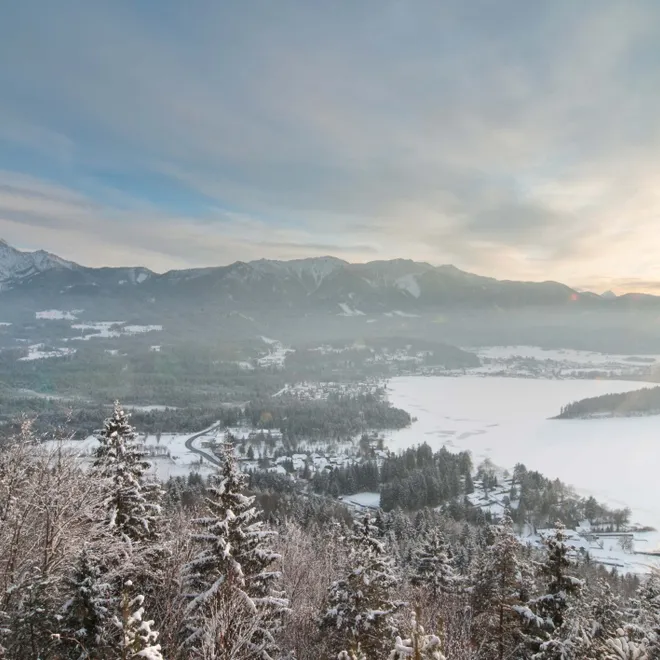 Winterlandschaft mit verschneitem Wald, zugefrorenem See und Sonnenaufgang über Bergkette