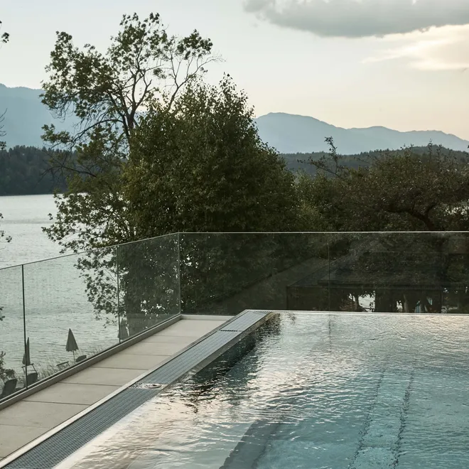 Infinity-Pool mit Glasgeländer mit Blick auf See und bewaldete Berge bei Sonnenuntergang