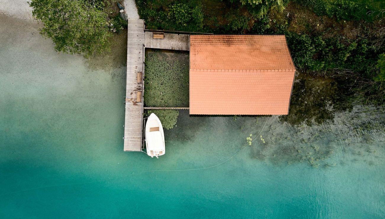Aerial view of a boat docked at a wooden pier next to a boathouse on turquoise lake