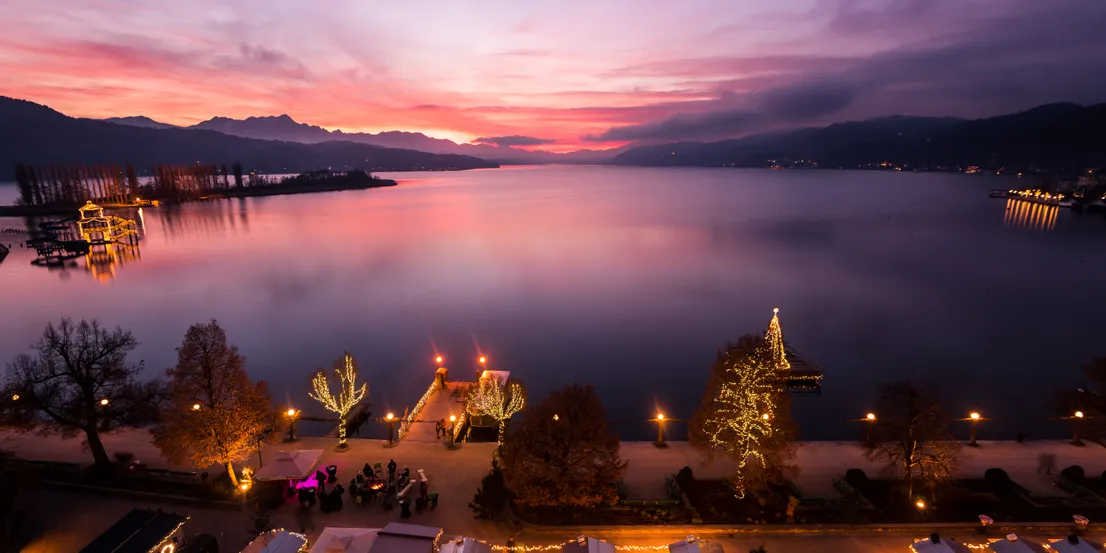 Weihnachtsmarkt am See bei Sonnenuntergang mit festlicher Beleuchtung und Bergkulisse