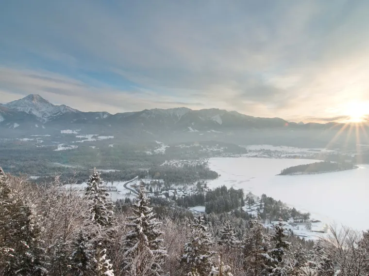 Winterlandschaft mit verschneitem Wald, zugefrorenem See und Sonnenaufgang über Bergkette