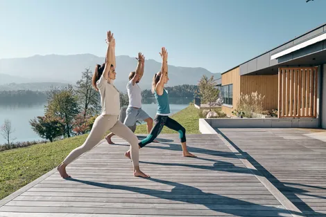Group practicing outdoor yoga on wooden deck near lake with mountain view