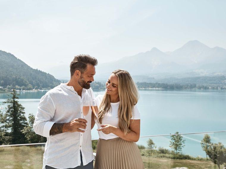Lächelndes Paar mit Weingläsern auf Balkon mit Blick auf Bergsee