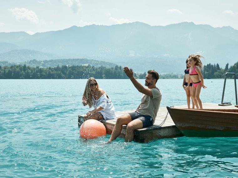 Familie spielt auf Holzsteg am Bergsee, während Kinder ins Wasser springen