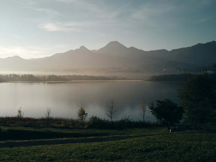 Ruhiger See mit nebligem Gebirge bei Sonnenaufgang und grünen Bäumen im Vordergrund