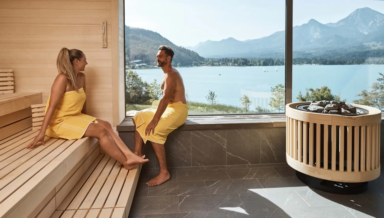 Paar entspannt in moderner Sauna mit Blick auf See und Berge durch großes Fenster