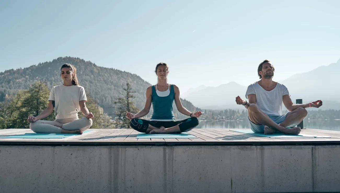 Drei Personen meditieren auf Yogamatten im Freien mit Blick auf einen Bergsee