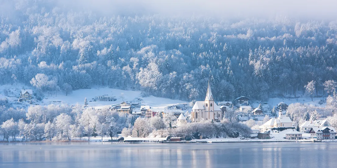 Schneebedecktes Dorf mit Kirche am See vor bewaldetem Berghang im Winter