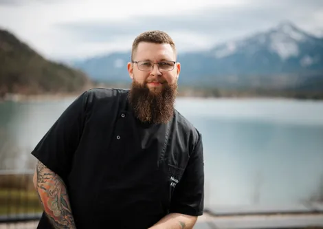 Bärtiger Koch in schwarzer Uniform posiert im Freien an einem See mit Bergblick