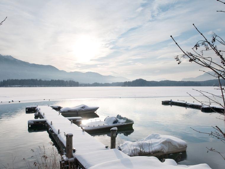 Verschneiter Holzsteg mit Booten auf zugefrorenem See in winterlicher Berglandschaft