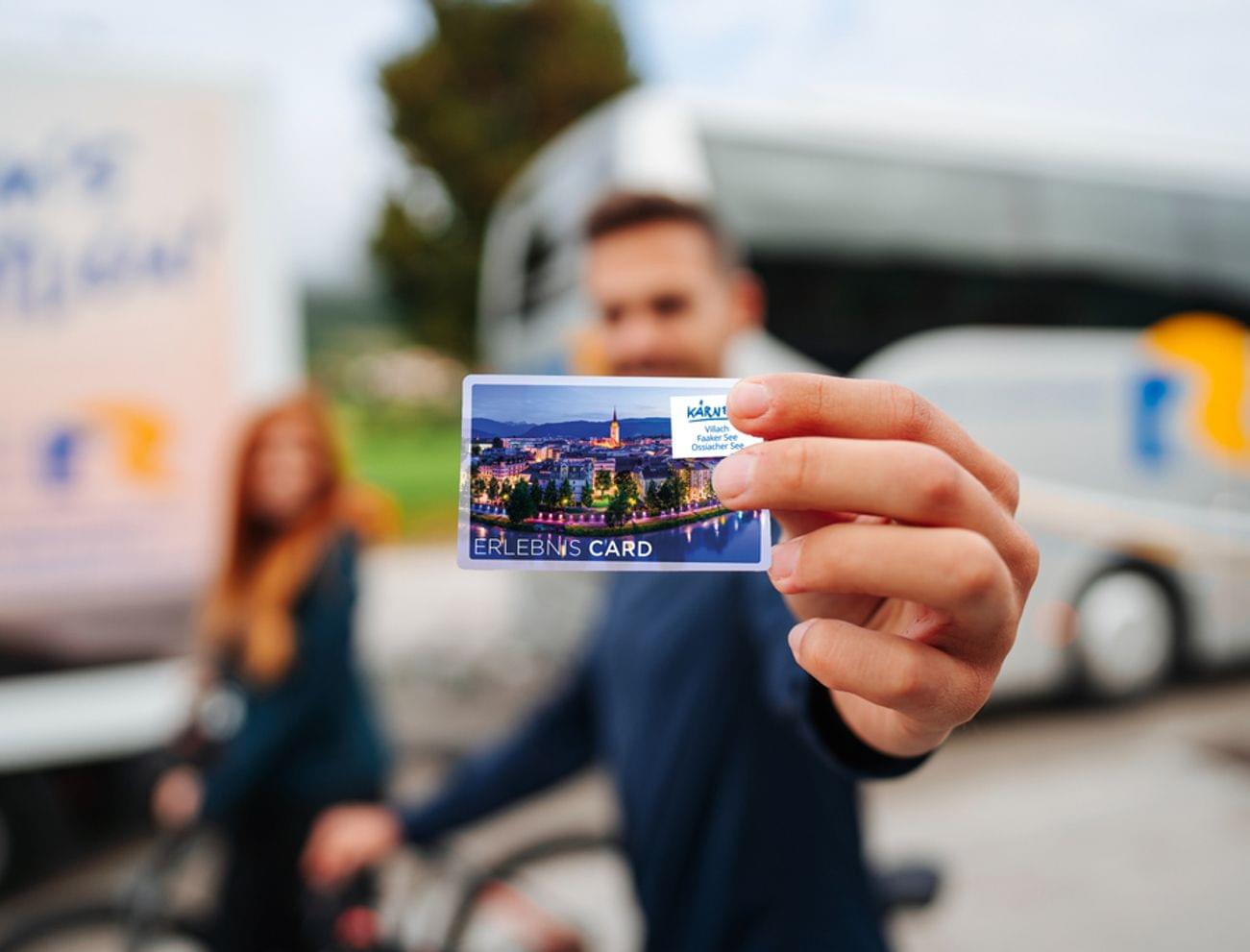 Mann hält Kärnten Erlebnis Card mit Stadtansicht vor einem Reisebus