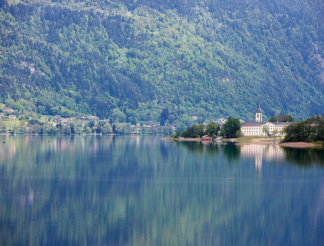 See mit Alpenort und Kirche, die sich im ruhigen Wasser vor bewaldeten Bergen spiegeln