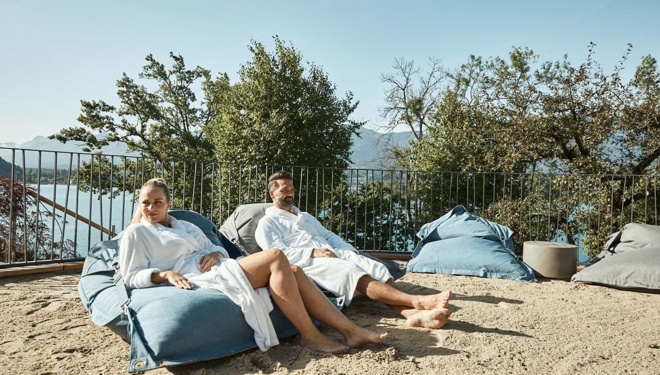 Paar entspannt im Bademantel auf Sitzsäcken auf sandiger Terrasse mit See- und Bergblick