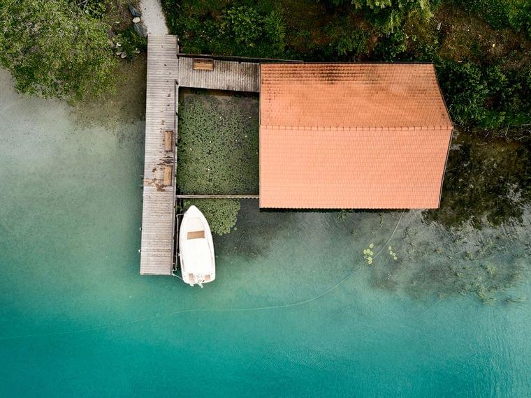 Aerial view of a boat docked at a wooden pier next to a boathouse on turquoise lake