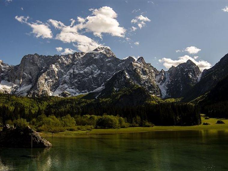 Catena montuosa innevata sopra una foresta verde e un lago alpino calmo sotto cielo blu
