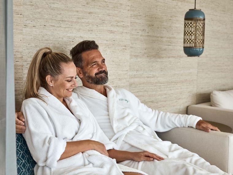 Smiling couple in white bathrobes relaxing on a spa lounge sofa