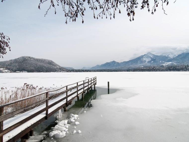 Verschneiter Holzsteg auf zugefrorenem Alpensee mit Blick auf verschneite Berge
