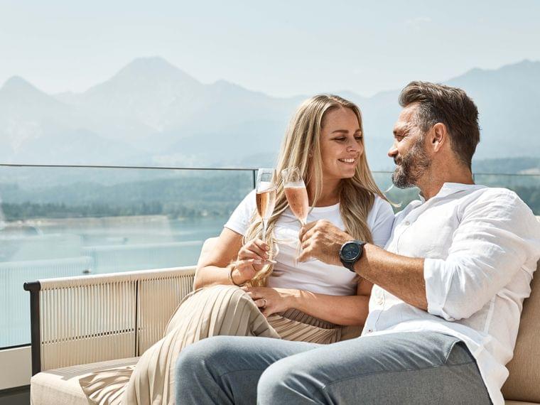 Coppia sorridente brinda con champagne rosé su terrazza soleggiata con vista lago di montagna
