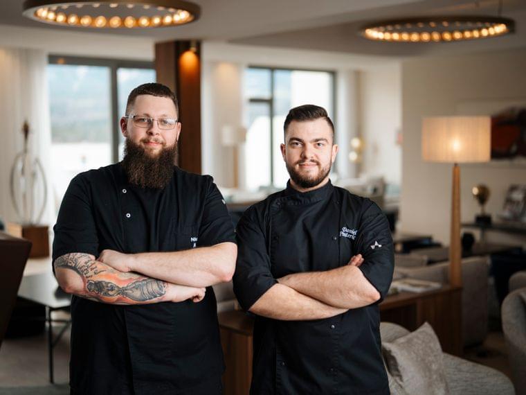 Zwei professionelle Köche in schwarzer Uniform posieren selbstbewusst in modernem Restaurantinterieur
