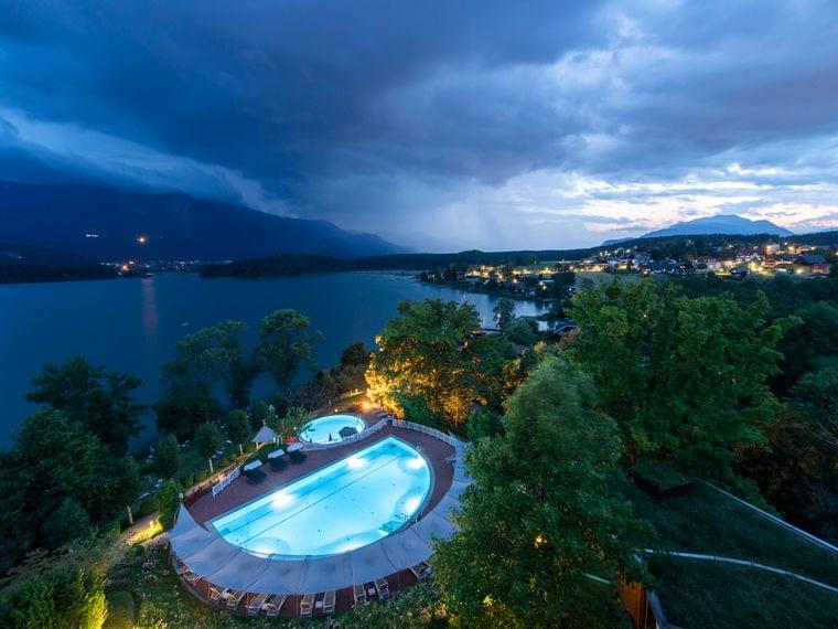 Luxury lakeside resort with illuminated pool and spa at dusk near mountain landscape
