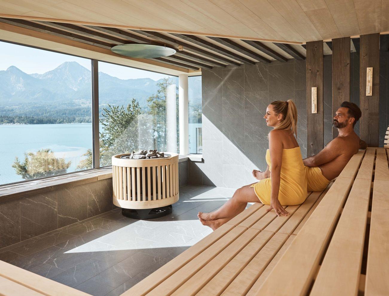 Couple relaxing in modern sauna with panoramic lake and mountain view