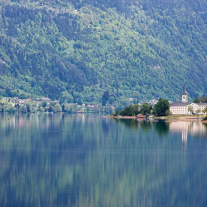 See mit Alpenort und Kirche, die sich im ruhigen Wasser vor bewaldeten Bergen spiegeln