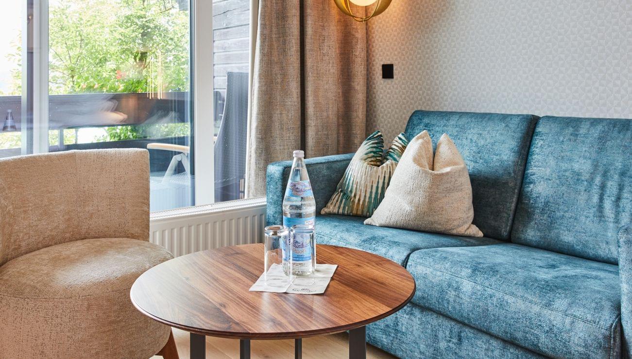 Modernes Hotelzimmer mit blauem Sofa, rundem Holztisch und Mineralwasser am Fenster
