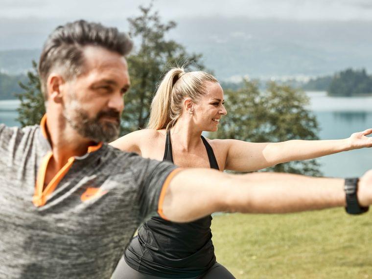 Man and woman doing outdoor yoga in warrior pose by a lake