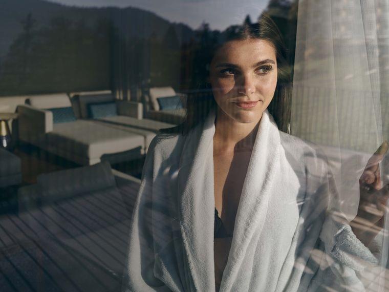 Donna in accappatoio bianco guarda fuori dalla finestra verso lounge rilassante del resort