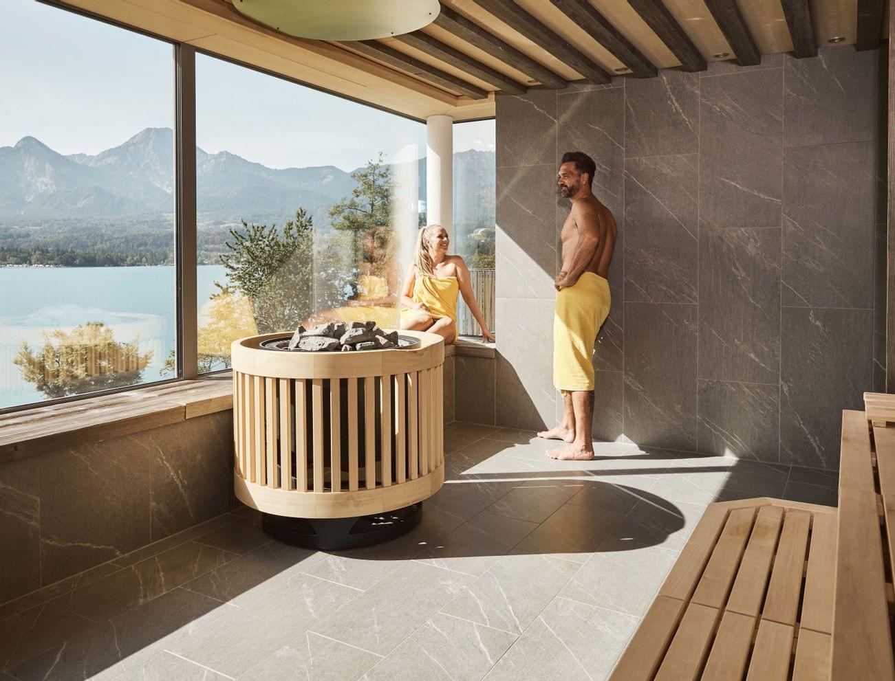 Couple relaxing in modern sauna with panoramic view of lake and mountains