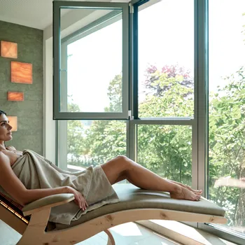 Frau entspannt auf Spa-Liege am großen Fenster mit Blick auf grüne Bäume