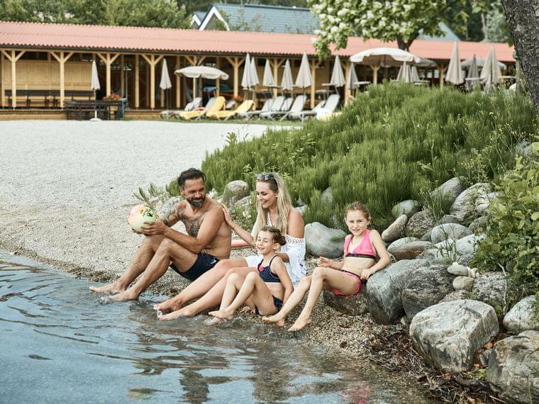 Glückliche Familie sitzt am Seeufer, Kinder planschen mit den Füßen im Wasser im See-Resort