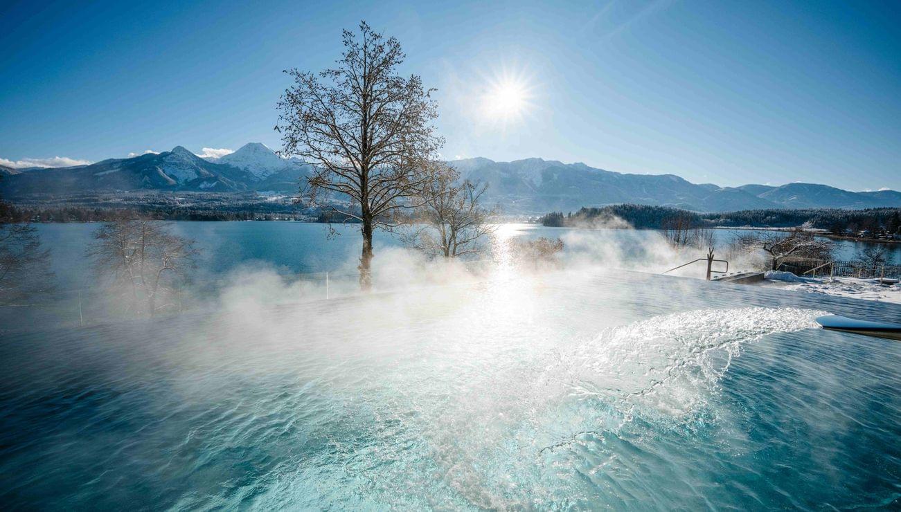 Piscina a sfioro fumante con vista su lago invernale e montagne innevate sotto cielo sereno