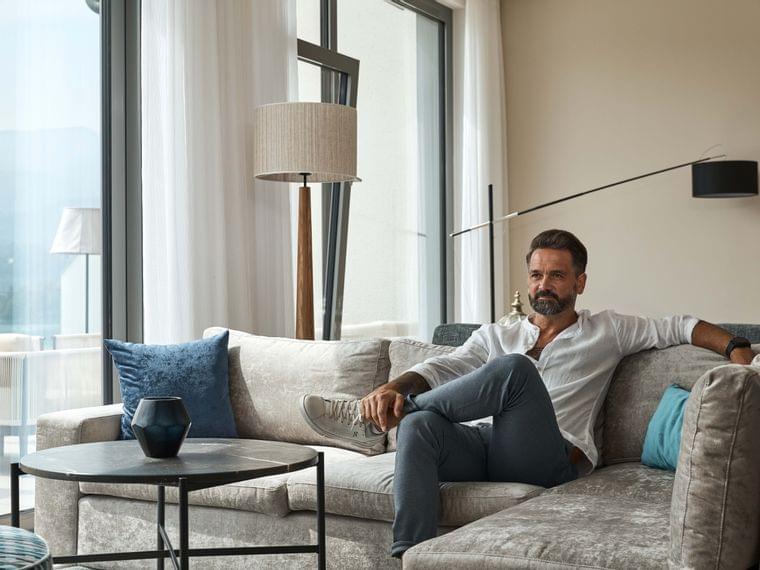 Man relaxing on modern grey sofa in bright living room with large windows